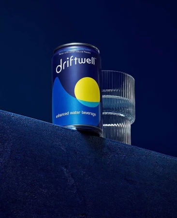 A single can of Driftwell enhanced water beverage is next to a clear glass on a dark blue textured surface against a dark blue background.