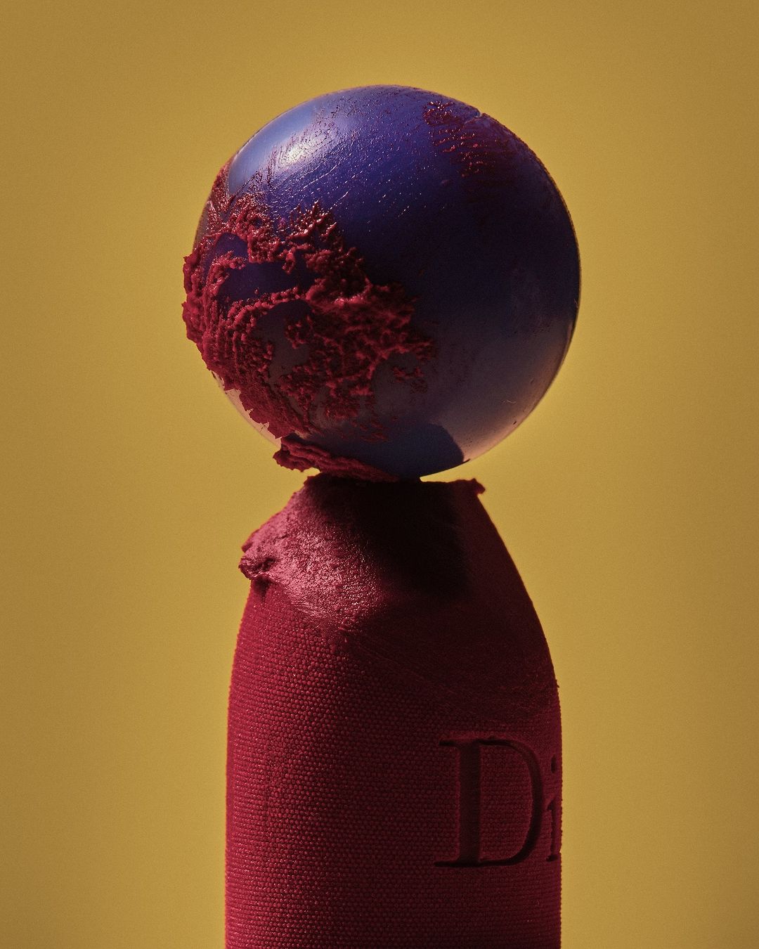 Macro studio shot of a blue spherical object partially coated with red, balanced on a red lipstick with a yellow studio backdrop.