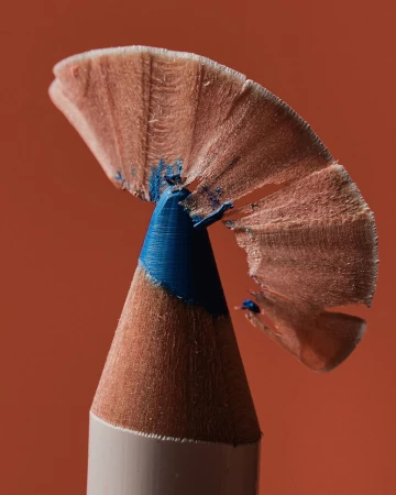 Close-up of a blue makeup eyeliner pencil with the wood shavings at the tip set against a solid orange backdrop.
