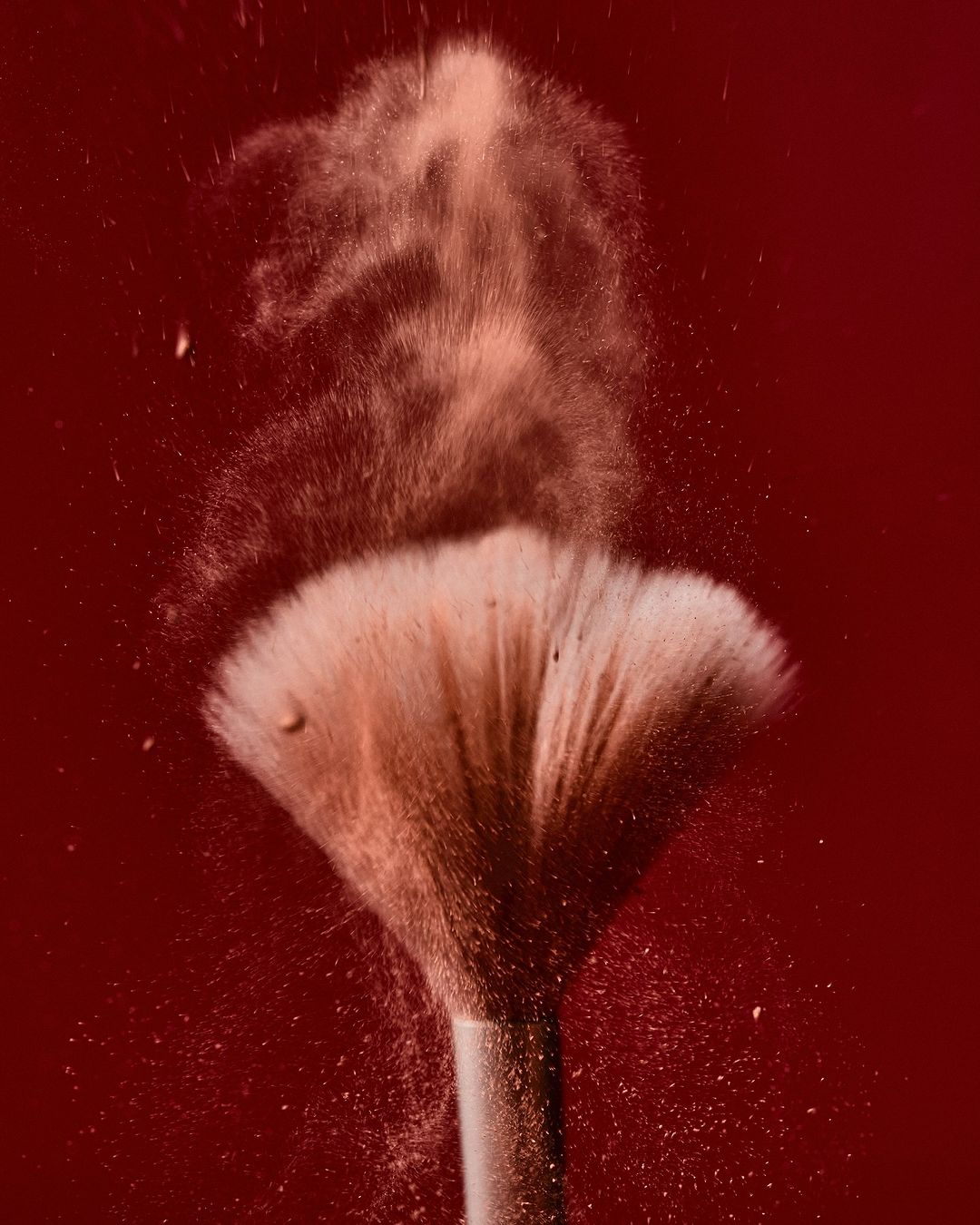 Close-up of a makeup brush with powder particles in the air against a dark red background, suitable for beauty product advertising.