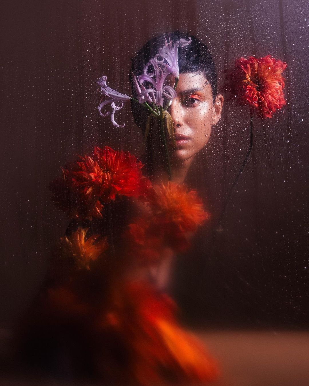 Portrait of a woman with vibrant floral accents and water droplets on glass, set against a softly lit, dark background.