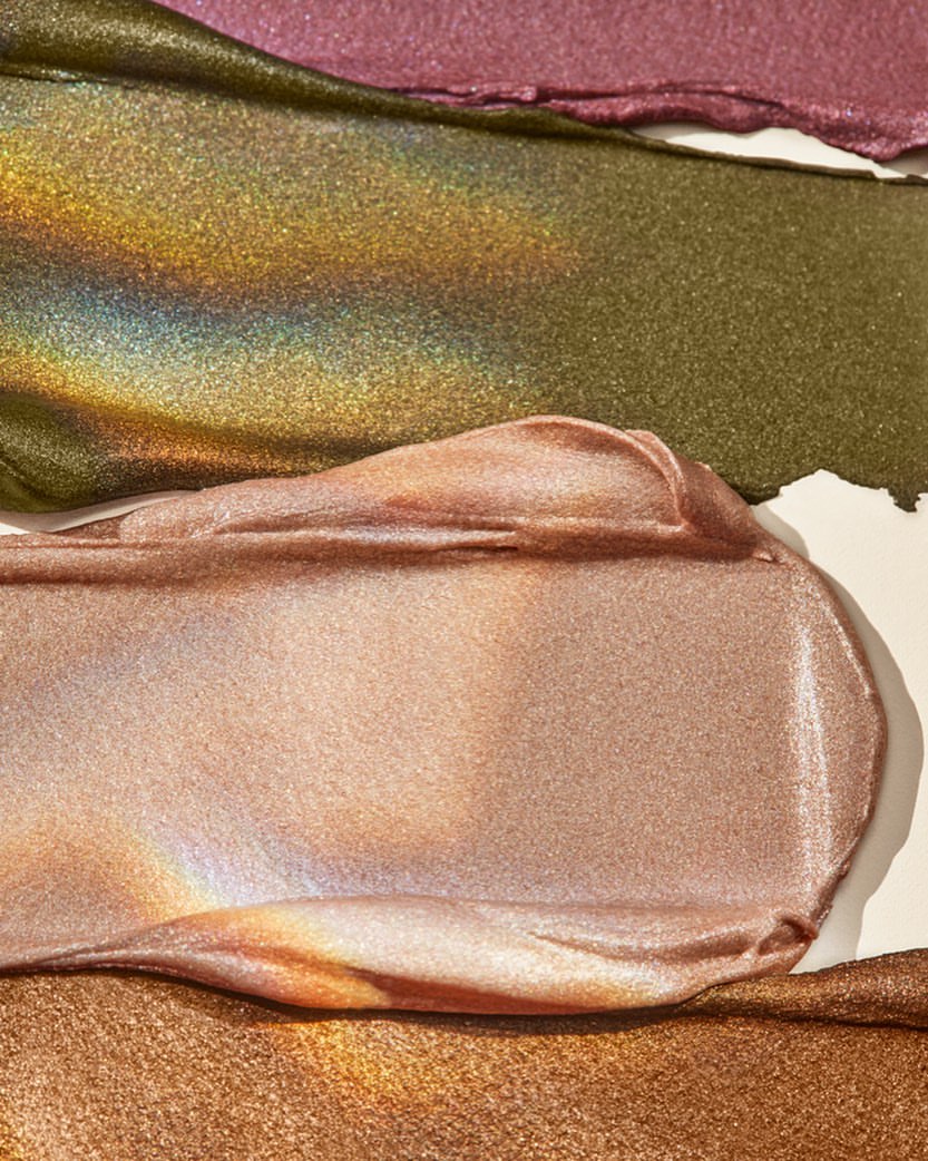 Close-up of textured smears of shimmering makeup in mauve, olive-gold, peach, and bronze shades against a neutral backdrop.
