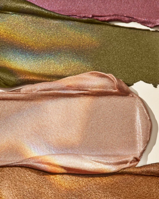 Close-up of textured smears of shimmering makeup in mauve, olive-gold, peach, and bronze shades against a neutral backdrop.