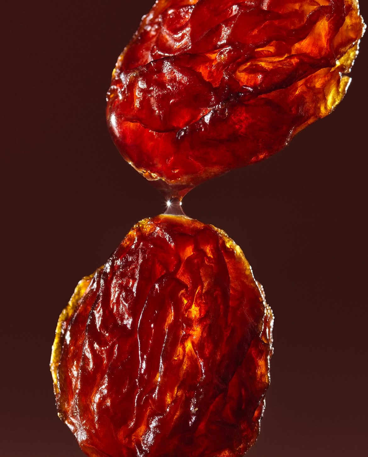 Close-up still life of two glistening, semi-translucent, reddish-brown lozenges connected by a droplet, set against a dark brown background.