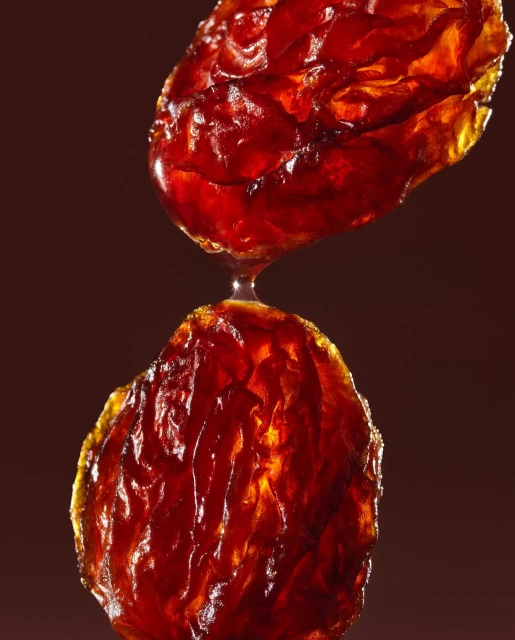 Close-up still life of two glistening, semi-translucent, reddish-brown lozenges connected by a droplet, set against a dark brown background.