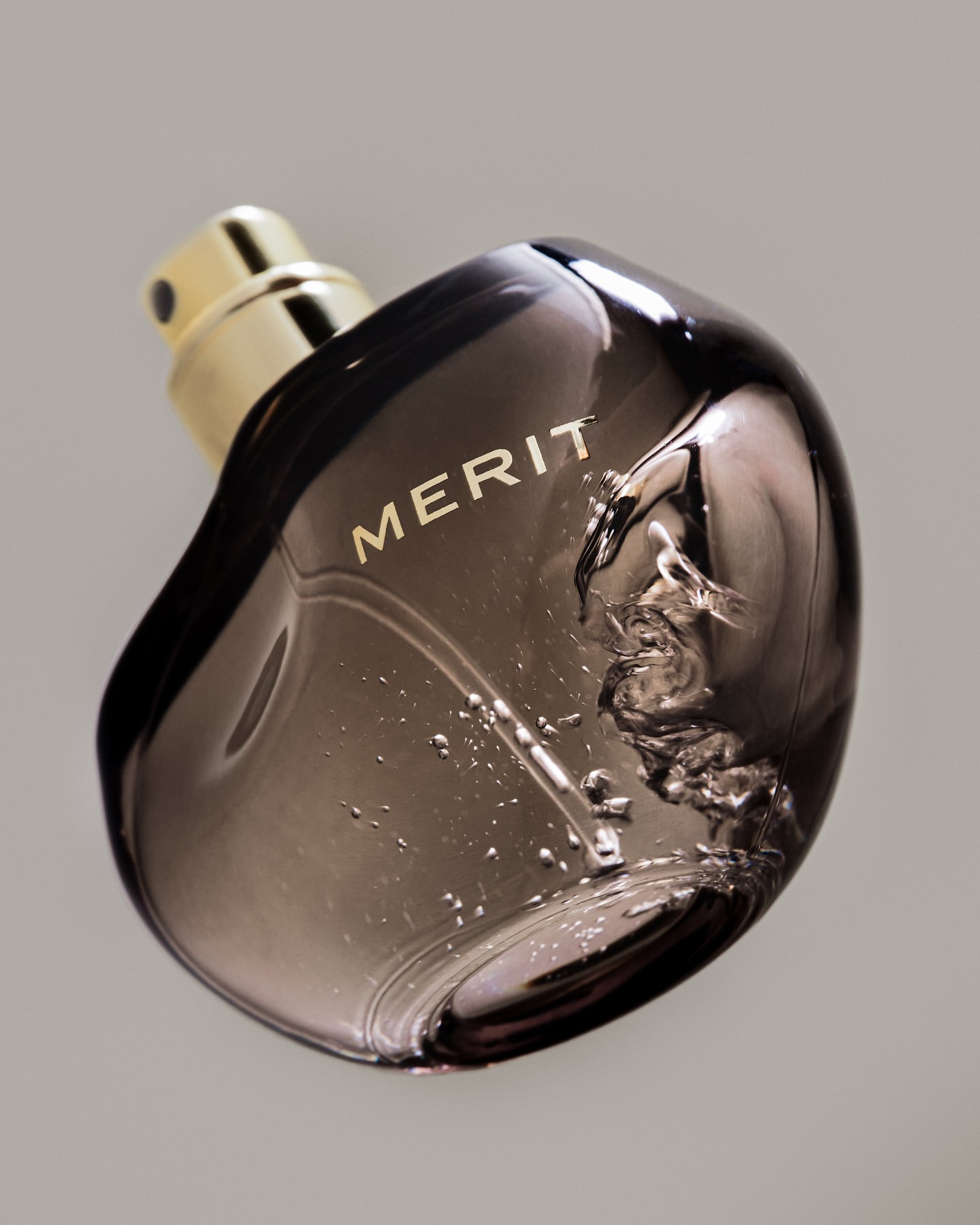 Studio shot of a dark glass perfume bottle with a gold nozzle and the word 'MERIT' on the front, against a neutral background.