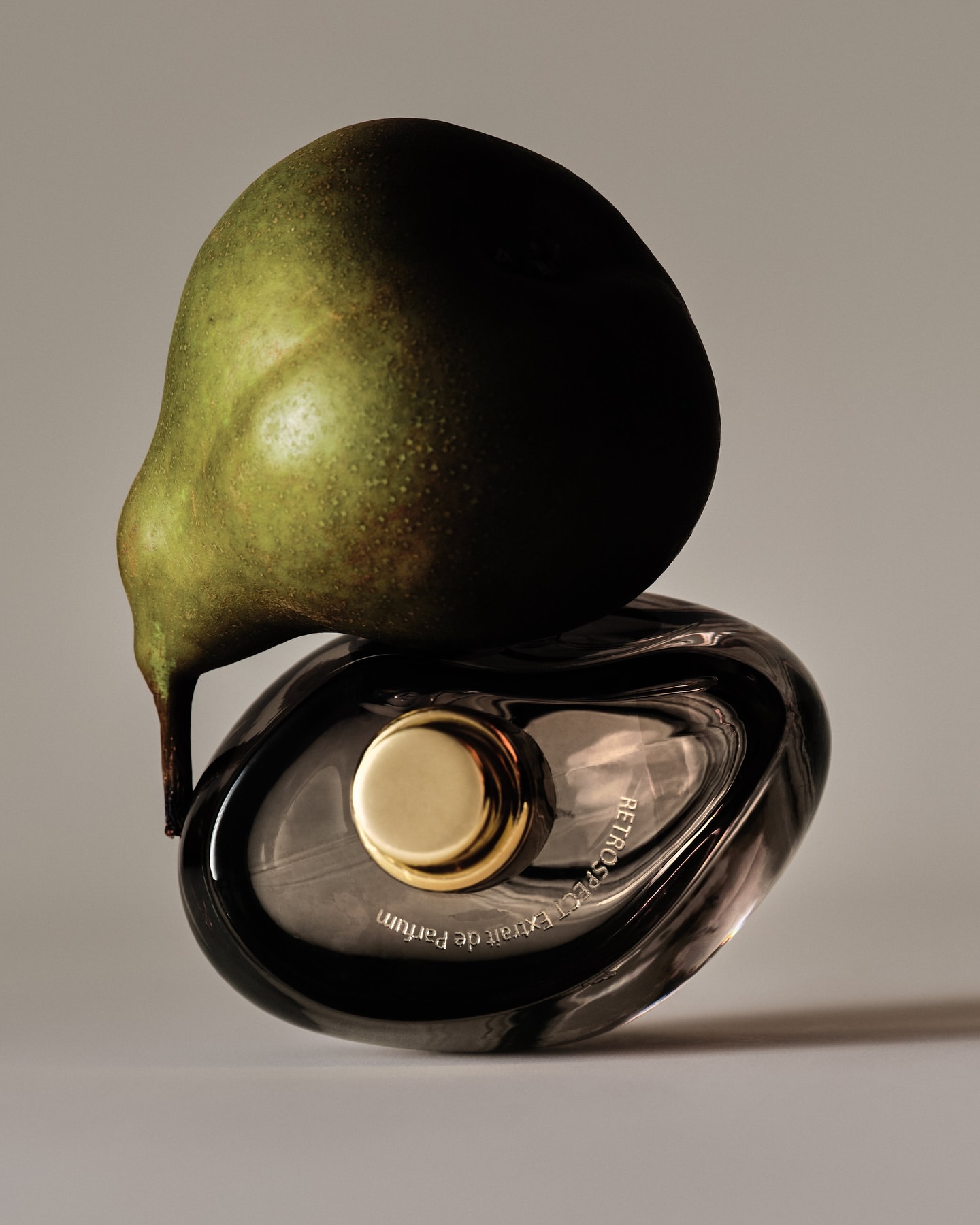 Still life of a green pear balancing on a gray glass perfume bottle with a gold cap on a grey background in a studio setting.