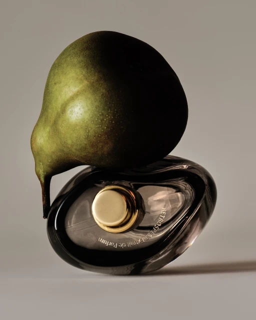 Still life of a green pear balancing on a gray glass perfume bottle with a gold cap on a grey background in a studio setting.