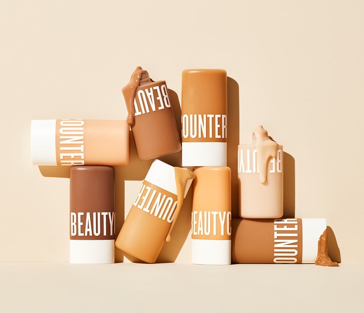 Several Beautycounter brand cylindrical containers of makeup foundation displayed at various angles, with beige monochrome background.
