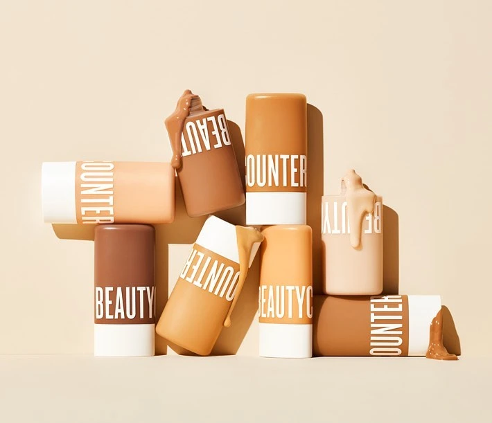 Several Beautycounter brand cylindrical containers of makeup foundation displayed at various angles, with beige monochrome background.