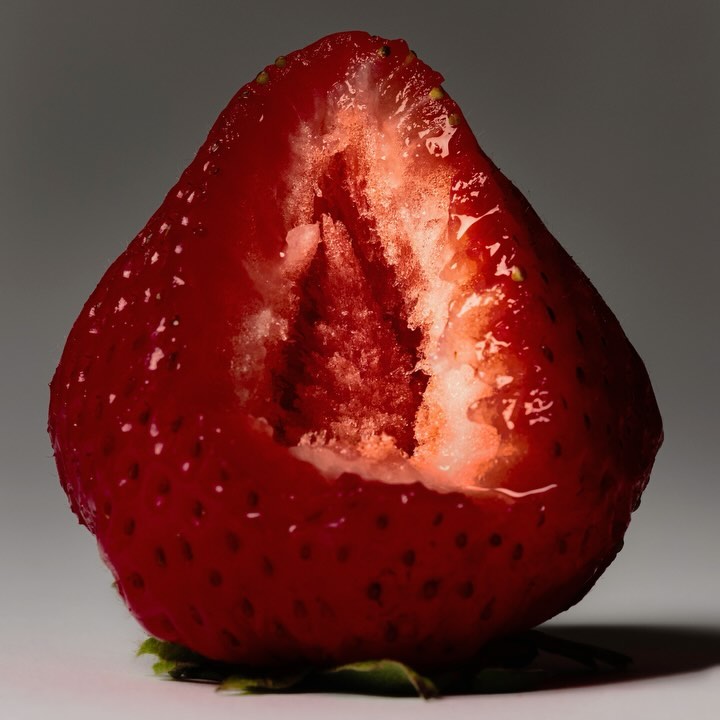Close-up shot of a strawberry cut in half. The vibrant red fruit contrasts with the grey background. The strawberry's seeds are visible.