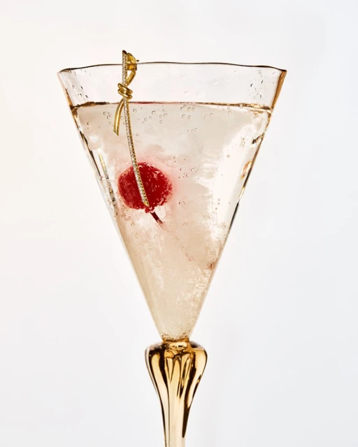 Elegant martini glass filled with a sparkling beverage, adorned with a cherry and a decorative gold cocktail pick on a clean white backdrop.