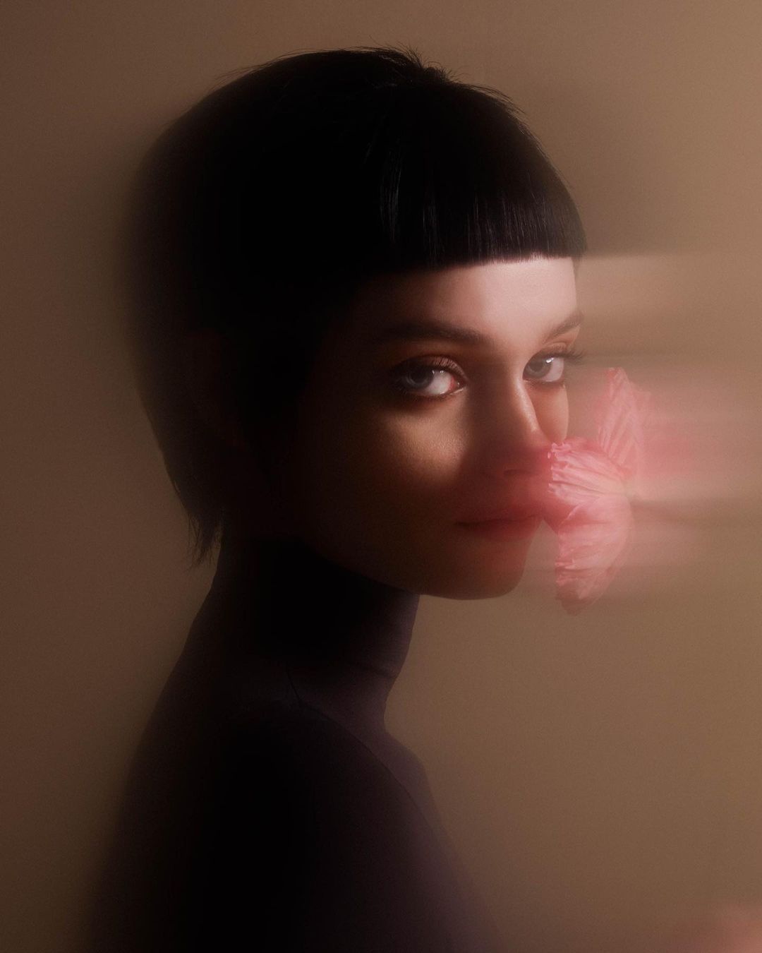 A young woman with short black hair poses indoors with a blurred pink flower near her face against a neutral-toned background.