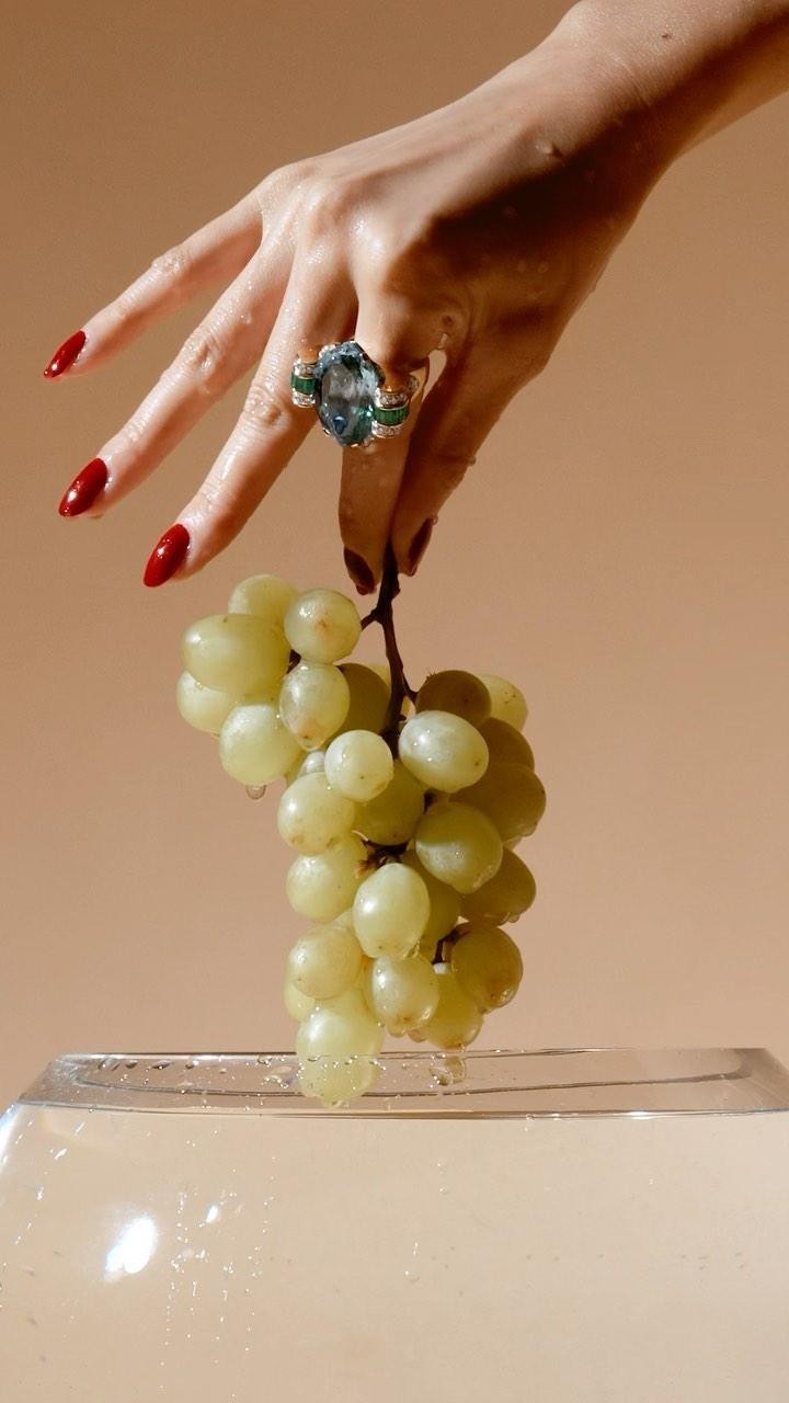 Hand with red nails holds green grapes over water with a large jewel ring on the index finger, with a tan background.