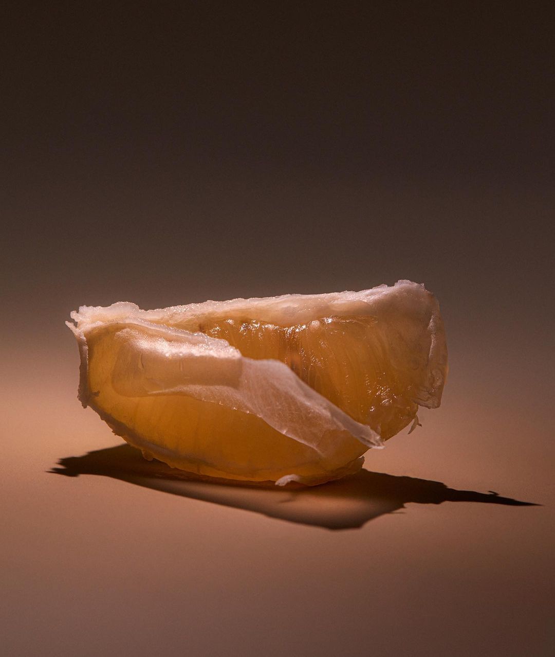 Close-up studio shot of a peeled orange slice casting a shadow on a flat surface with a soft, diffused brown-toned background.