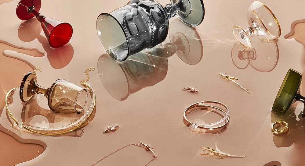 Arrangement of tipped wine glasses and gold jewellery spread across a glossy surface with liquid spills and visible reflections.