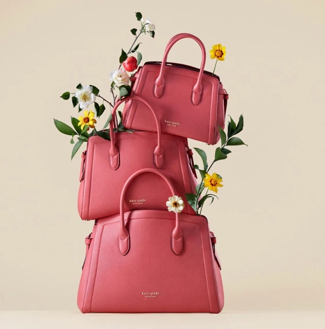Three Kate Spade purses stacked against a neutral background, accented with sprigs of flowers in white, yellow, and green.