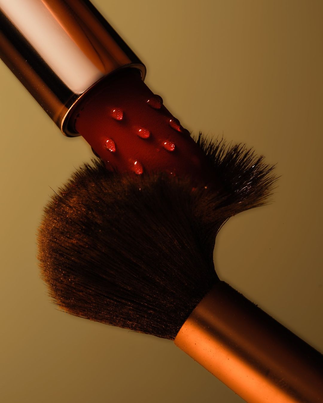 A close-up shot captures a makeup brush with dark bristles and a metallic handle, juxtaposed with a lipstick covered in water droplets.