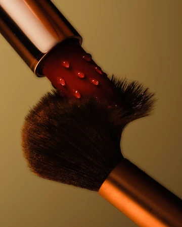 A close-up shot captures a makeup brush with dark bristles and a metallic handle, juxtaposed with a lipstick covered in water droplets.