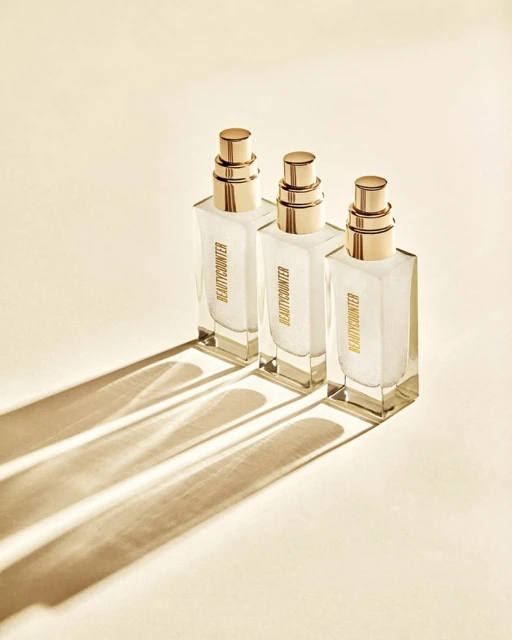 Three square glass bottles with gold pumps stand in a row casting shadows on a cream surface, showcasing minimalist beauty product design.