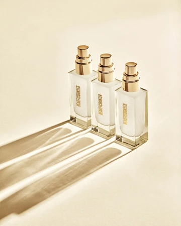 Three square glass bottles with gold pumps stand in a row casting shadows on a cream surface, showcasing minimalist beauty product design.
