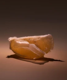 A close up studio photograph of a single translucent citrus segment resting on a smooth neutral surface.
