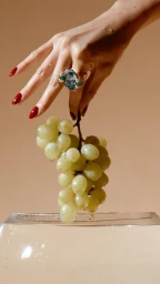 Close-up of a hand with red nail polish holding a bunch of green grapes above a glass container against a warm beige background.