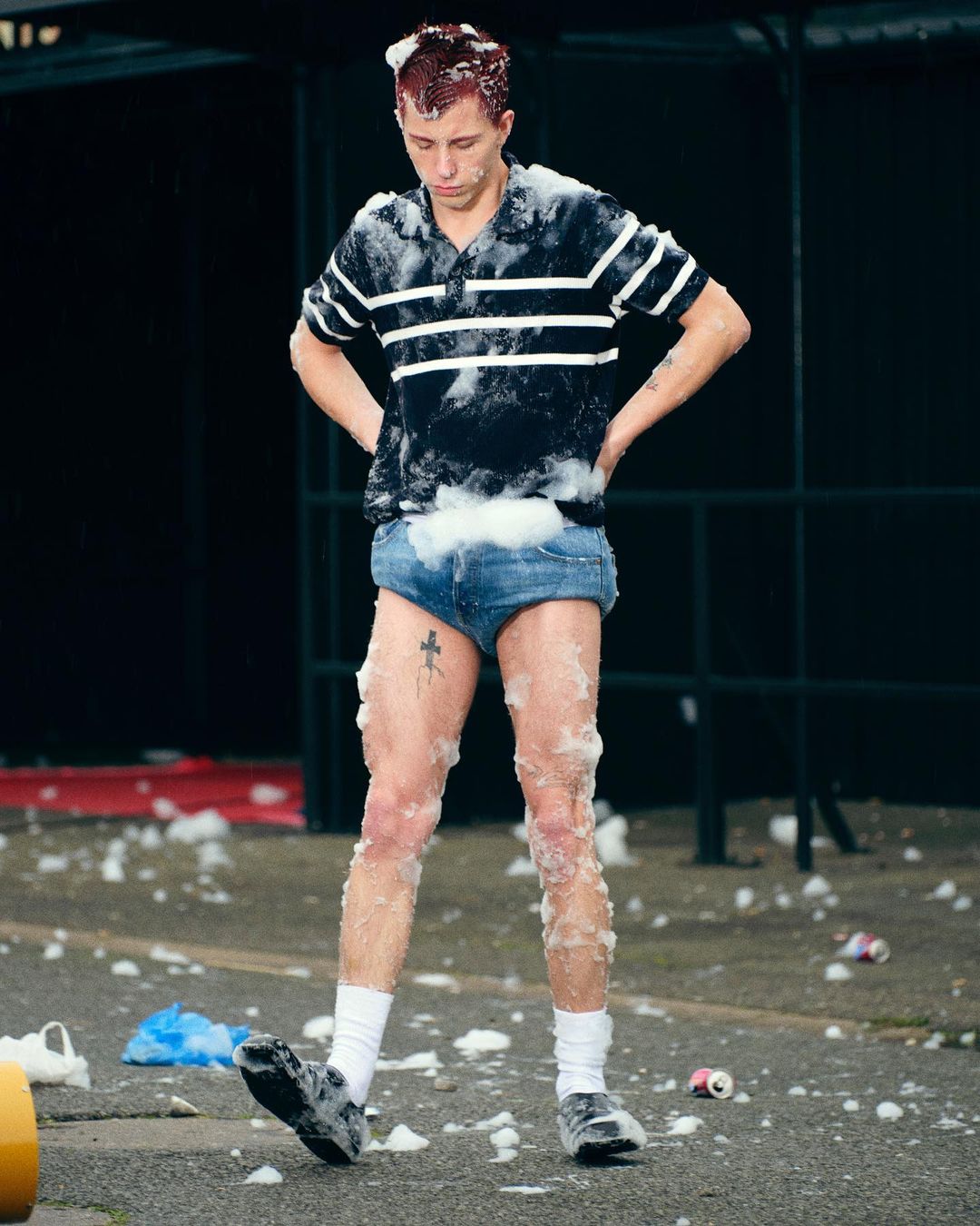 Young adult male stands covered in foam with hands on hips, wearing shorts, socks, and shoes, with a black background and litter on the ground.