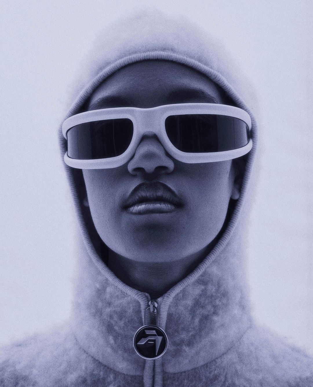Close-up portrait of an adult wearing white futuristic sunglasses and a hooded top against a light background, minimalist fashion.