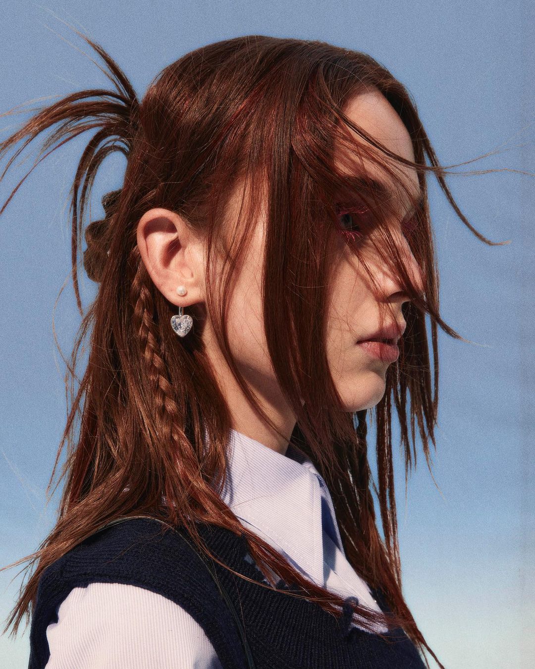 Young woman with auburn hair wearing a blue vest and heart-shaped earrings posing outdoors against a blue sky background.