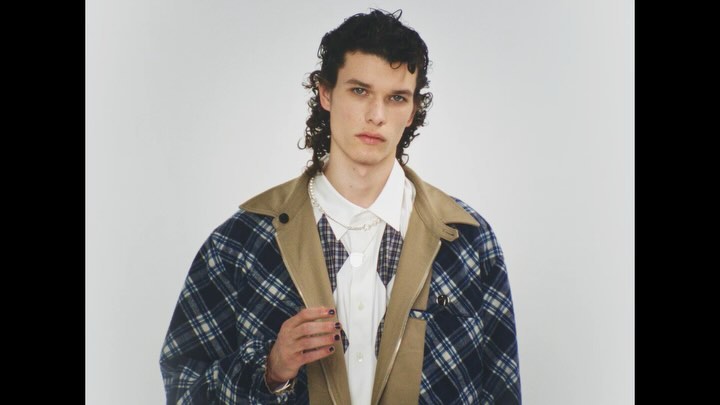 Adult male with curly hair, wearing a plaid jacket over a white shirt and vest, poses indoors against a white backdrop.
