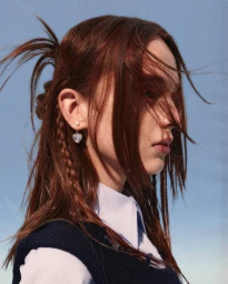 Profile portrait of a woman with red hair wearing heart-shaped earrings against a clear blue sky background.