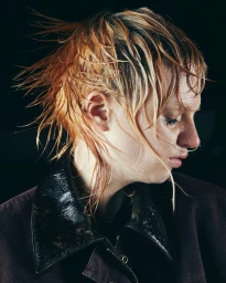 Profile portrait of a woman with wet, spiky blonde hair wearing a dark jacket against a black background by Suzie And Leo.