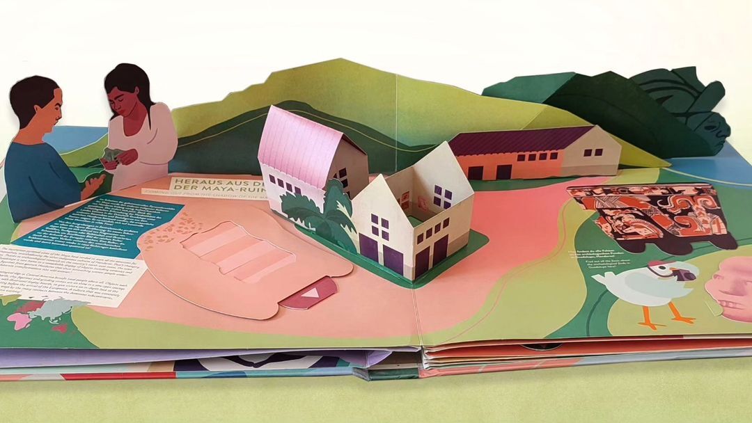 Pop-up book featuring two paper figures, paper houses, and a paper landscape with a mountain range, set against a white background.