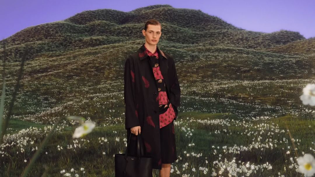 Male model poses in a black coat with red accents and a matching shirt and shorts in a field with rolling hills in the background.