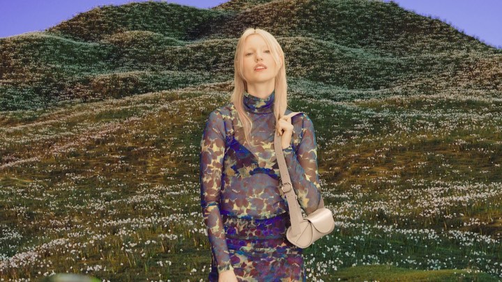 Woman in floral top and skirt with a purse standing in a field of flowers against a rolling hill landscape on a sunny day.
