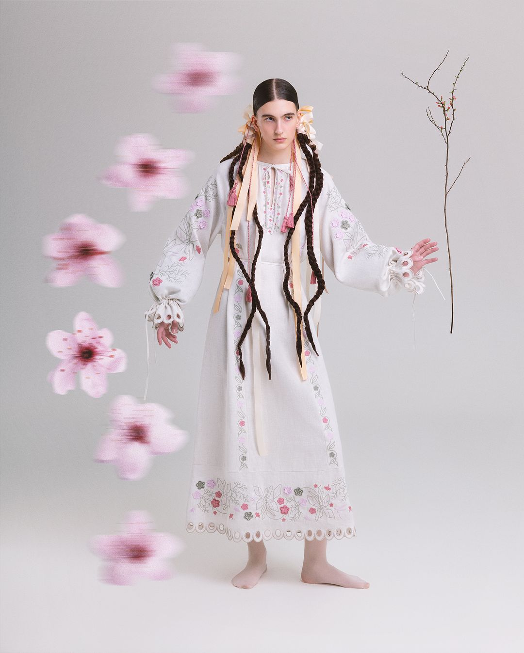 Woman in white embroidered dress with braids, standing in studio with blurred pink flower petals and branch. Soft, neutral lighting.