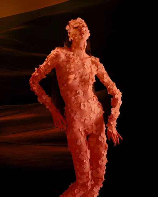Figure of a woman covered in peach-toned flower-like material, posing against a dark backdrop with orange highlights, fashion photography.