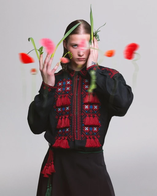 Portrait of a person in an embroidered black shirt surrounded by out-of-focus flowers in a studio setting, showcasing fashion and beauty.