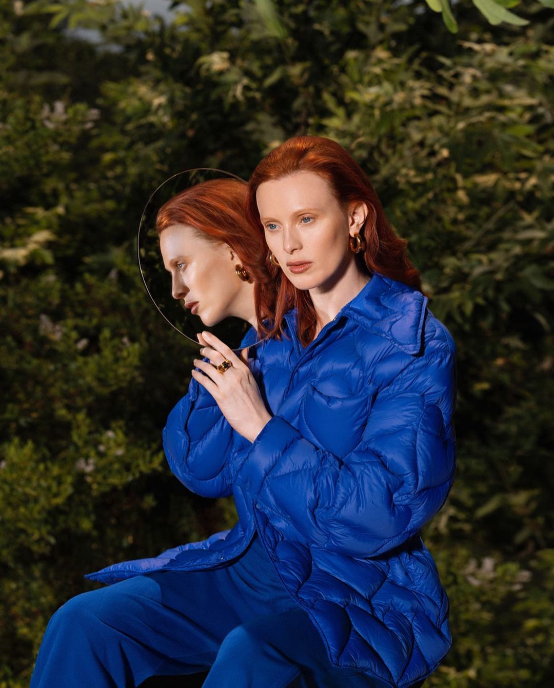 Redhead woman in blue quilted jacket, sitting with reflection in round mirror amidst green foliage. Fashion and nature blend seamlessly.