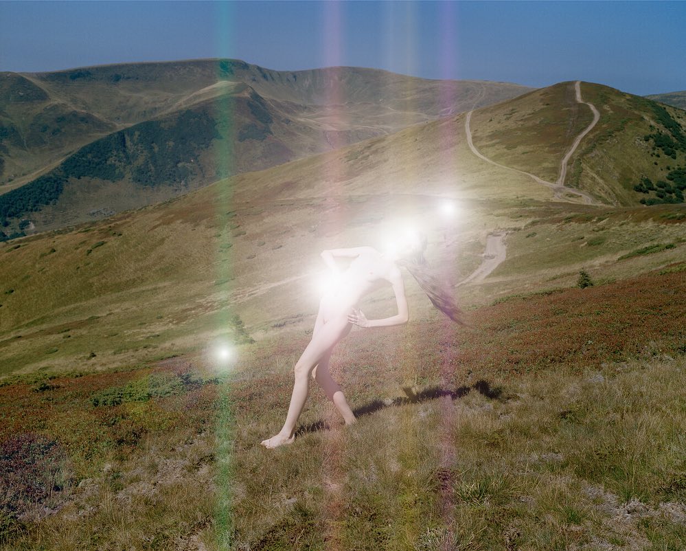 Abstract outdoor portrait of a bright figure standing in a grassy, hilly landscape under a clear blue sky. Lens flare creates glowing effects.