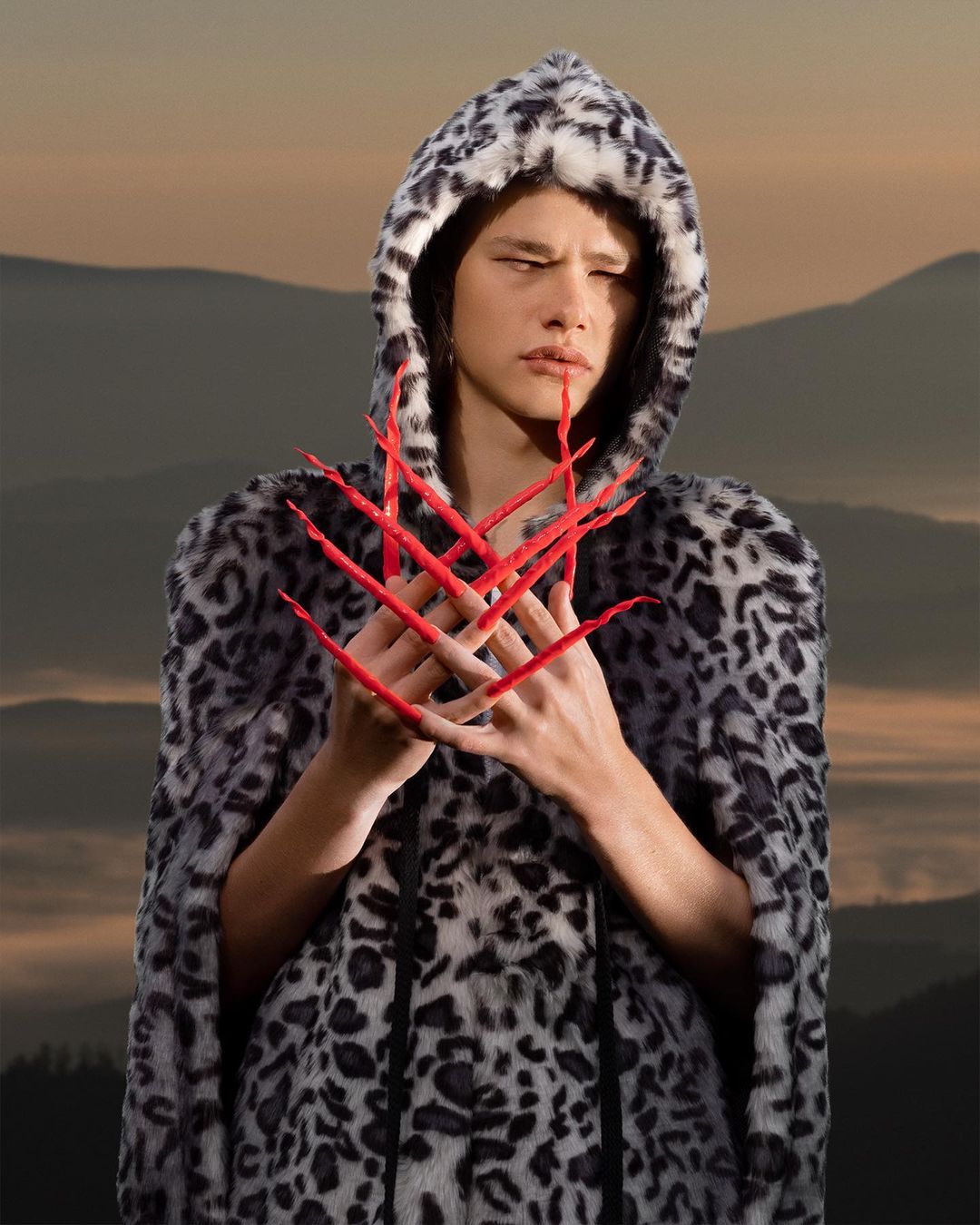 Portrait of a person with long red fingernails wearing a hooded jacket with leopard print, posing in front of a mountain landscape.