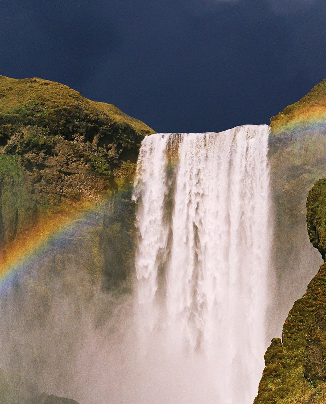 A picturesque waterfall cascades down mossy cliffs under a dark sky, creating a vibrant rainbow in the misty air near lush green cliffs.
