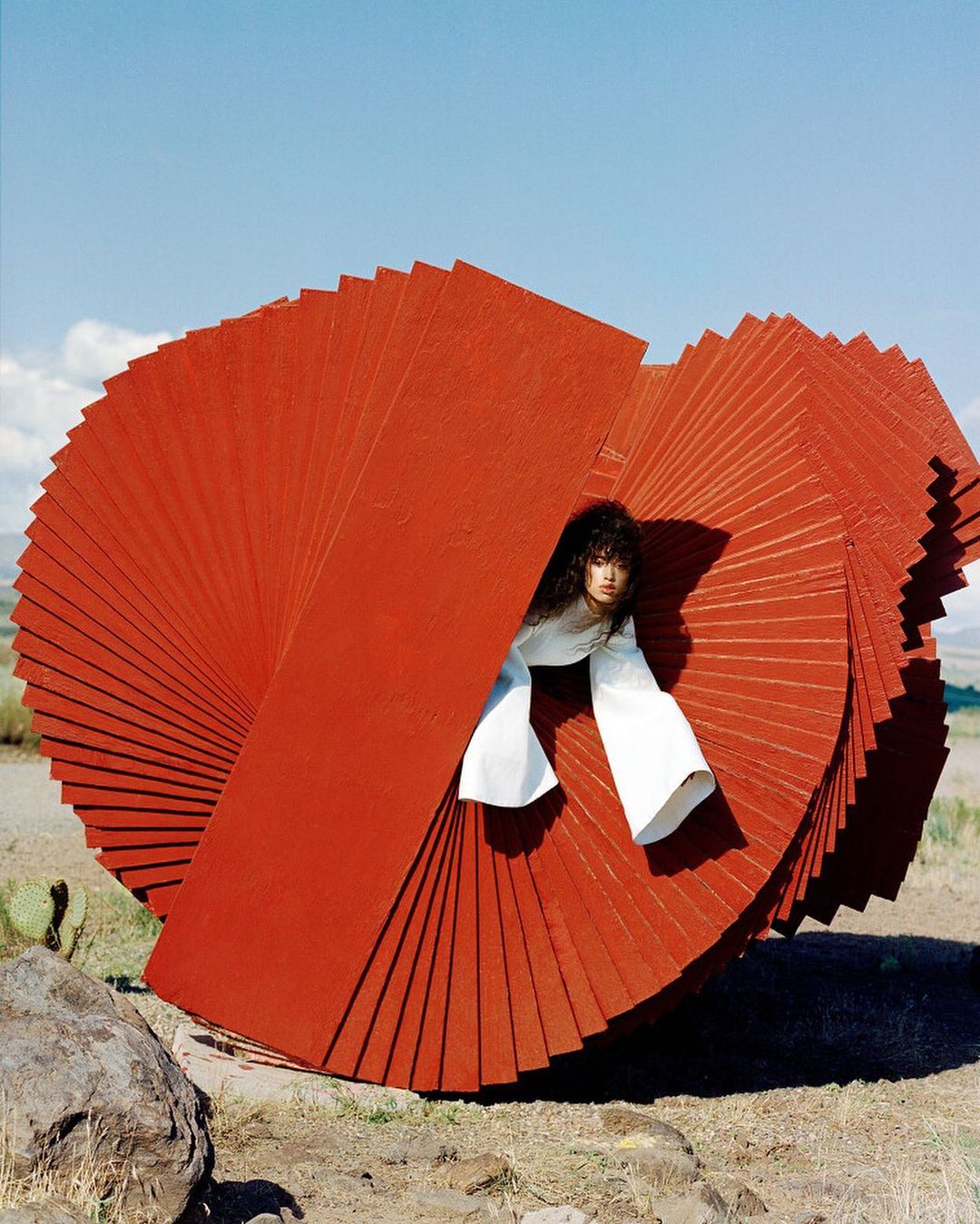 A woman poses next to a large, rust-colored sculpture in a field, wearing a white top with bell sleeves, under a clear blue sky.
