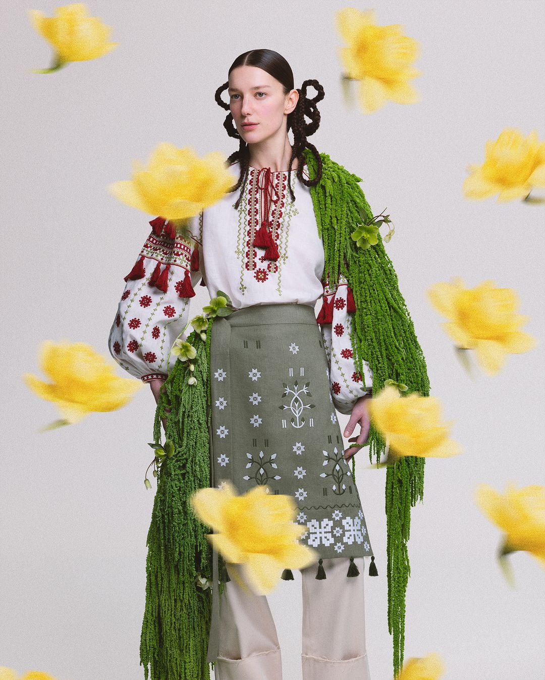 A woman in traditional clothing stands against a plain backdrop, surrounded by floating yellow flowers in a studio setting.