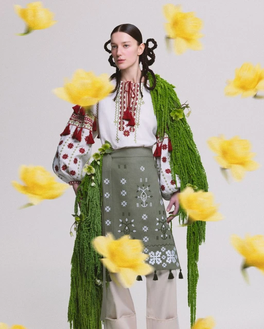 A woman in traditional clothing stands against a plain backdrop, surrounded by floating yellow flowers in a studio setting.