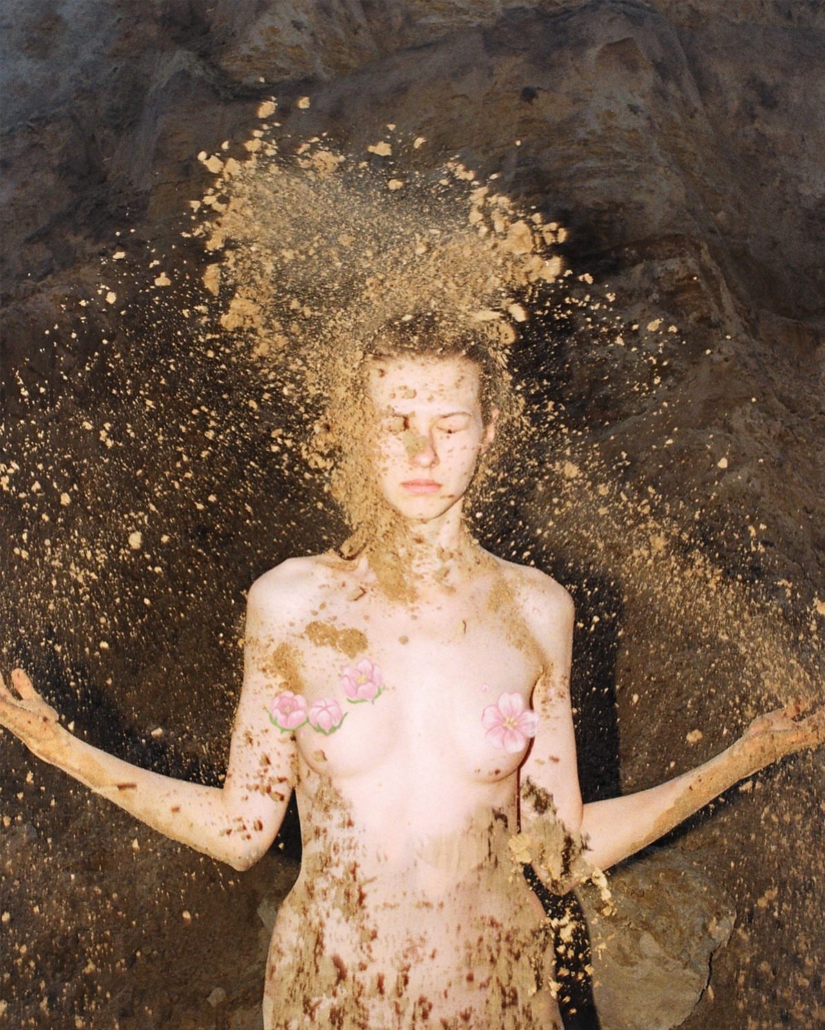 Woman covered in sand stands with arms outstretched in a cave. The scene is illuminated by natural light, highlighting textures.