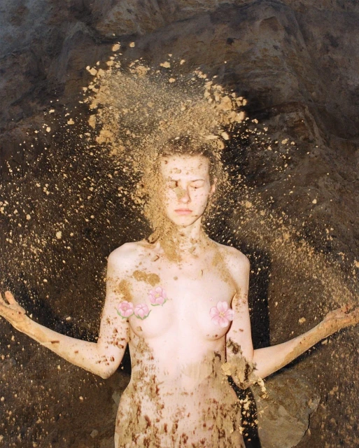 Woman covered in sand stands with arms outstretched in a cave. The scene is illuminated by natural light, highlighting textures.
