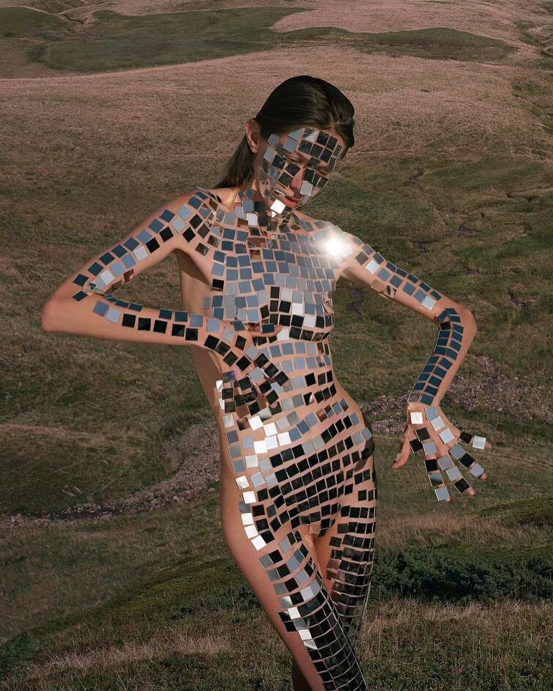 Adult female model covered in square mirror tiles posing in an open grassy field with rolling hills under overcast sky. 