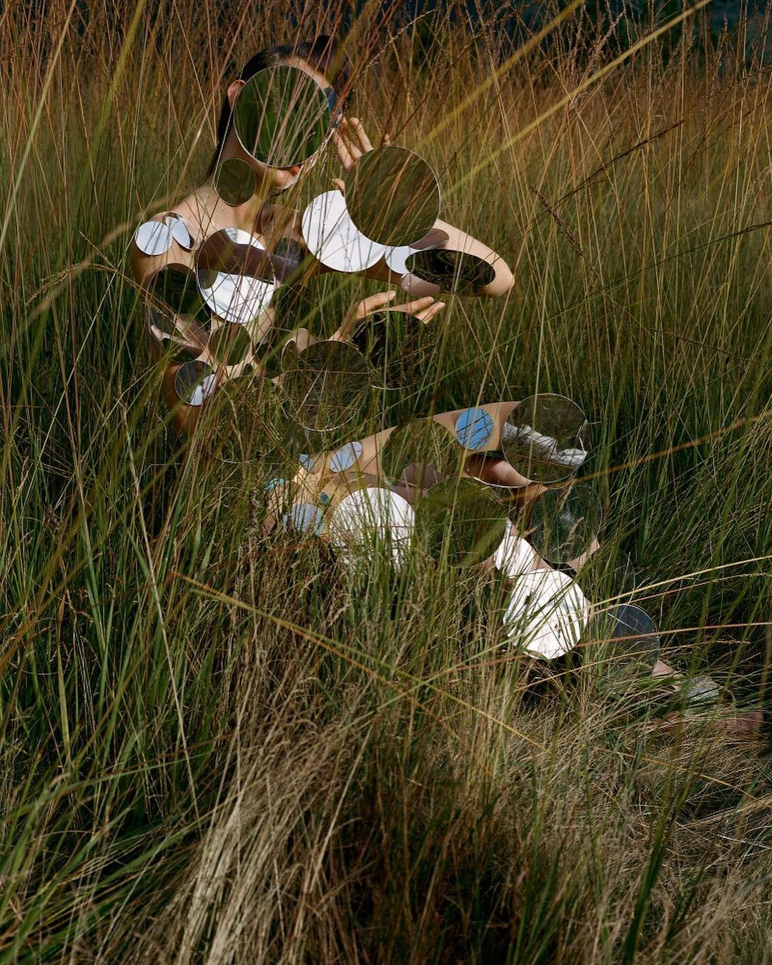 Surreal image of a fair-skinned person partially hidden in tall grass, featuring reflective circular objects obscuring the face and body.